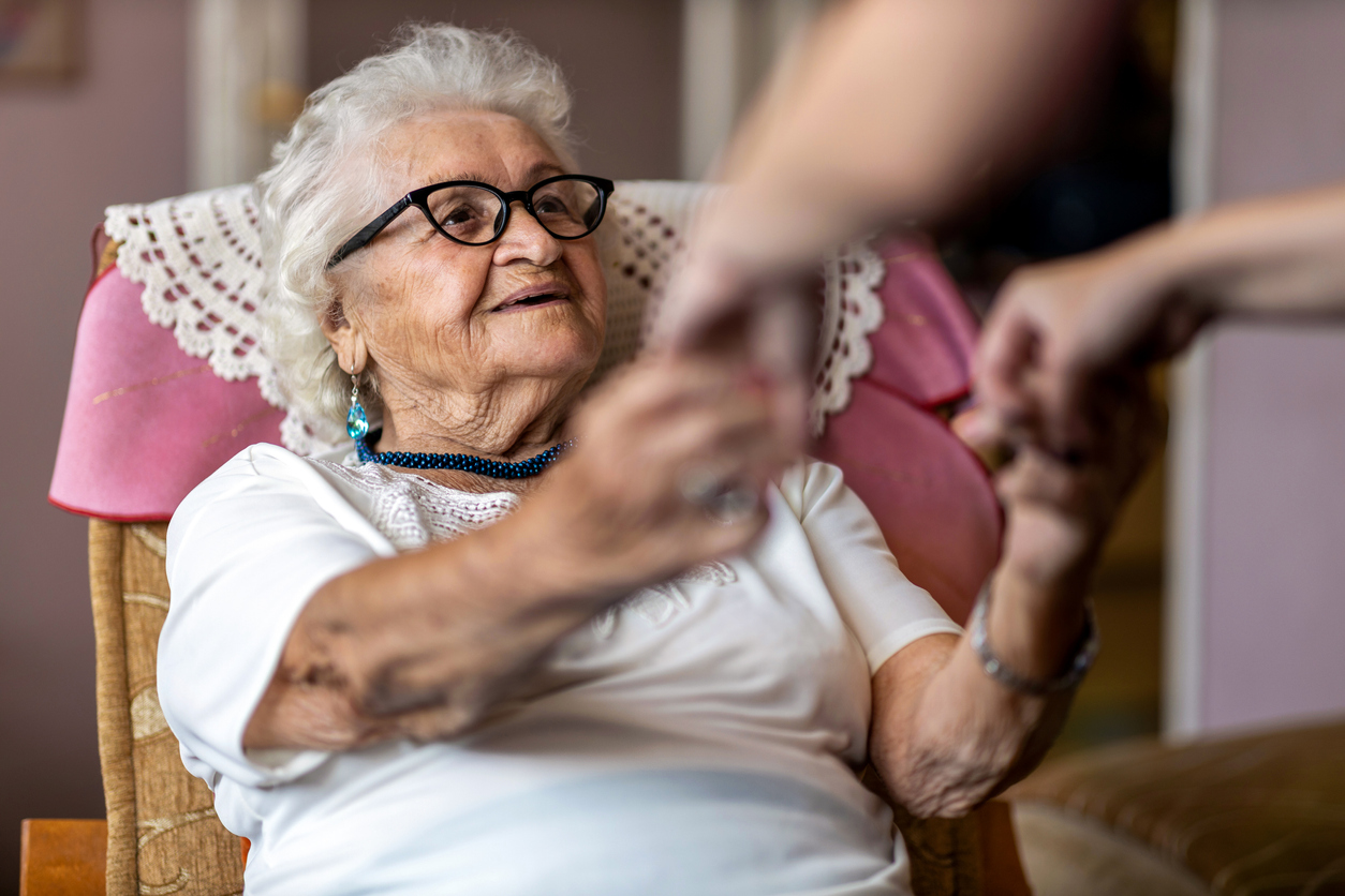 Female home caregiver supporting old woman to stand up from the armchair at care home